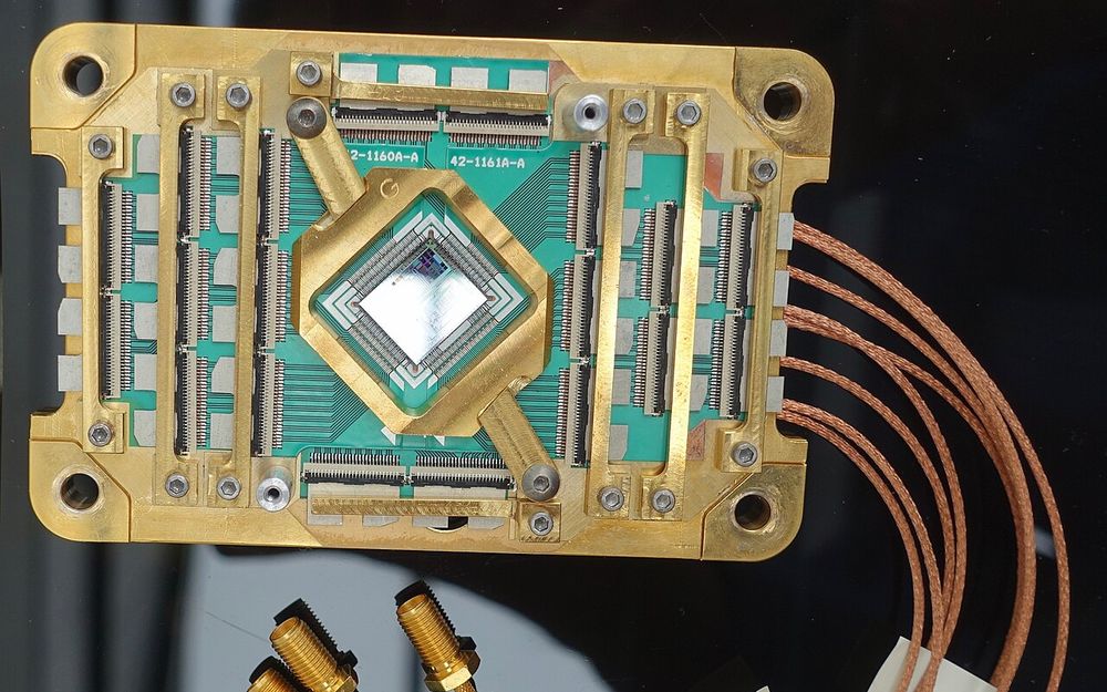 A square silicon chip at the centre of a gold-plated mounting board with green circuit traces and braided copper cables.