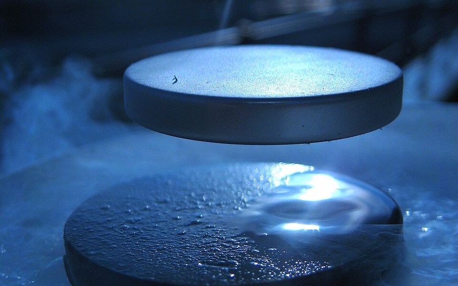 A small disc-shaped magnet hovering above a dark, frosted superconducting disc under blue light.