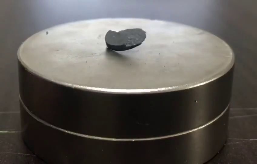 A small dark fragment of LK-99 tilted above a large disc-shaped magnet.